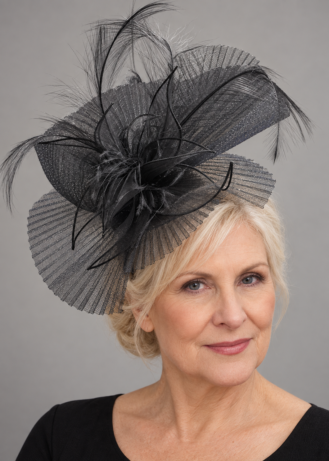 Mature blonde model wearing a large black pleated crinoline racewear fascinator with feather accents on an Alice band.