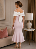 Back view of light blush pink midi mother of the bride dress featuring a dramatic ivory bow across the shoulders, pearl-beaded detailing, fitted crepe silhouette and fluted hem, modelled by a mature brunette woman in an elegant indoor boutique setting.