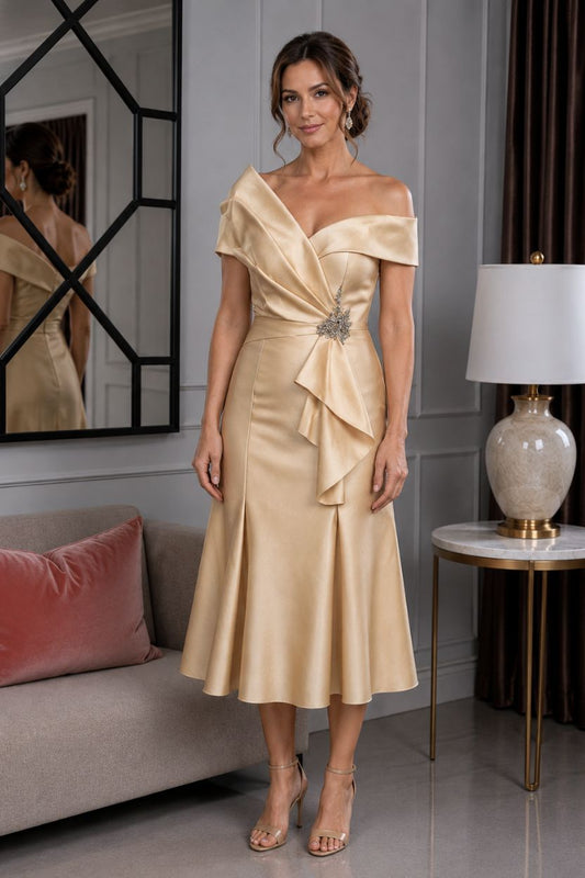 Cream gold off-the-shoulder satin mother of the bride dress with draped bodice detail and embellished waist, shown on a brunette size 12/14 model in an elegant indoor boutique setting.