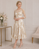 Mother of the bride fit-and-flare dress in ivory and gold jacquard with illusion sleeves and full midi skirt, styled for a formal wedding.