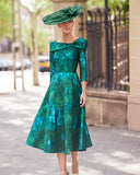 Elegant green brocade fit and flare dress with floral pattern, worn by a woman with a large coordinating hat, standing in an outdoor urban setting.