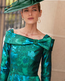 Elegant off-the-shoulder green floral brocade dress with a decorative bow detail, paired with a large green hat and dangling earrings.