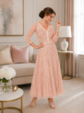 Blush pink embellished tea-length occasion dress with sheer three-quarter sleeves, fitted bodice and flowing A-line skirt, styled for mother of the bride or mother of the groom in an elegant indoor boutique setting.