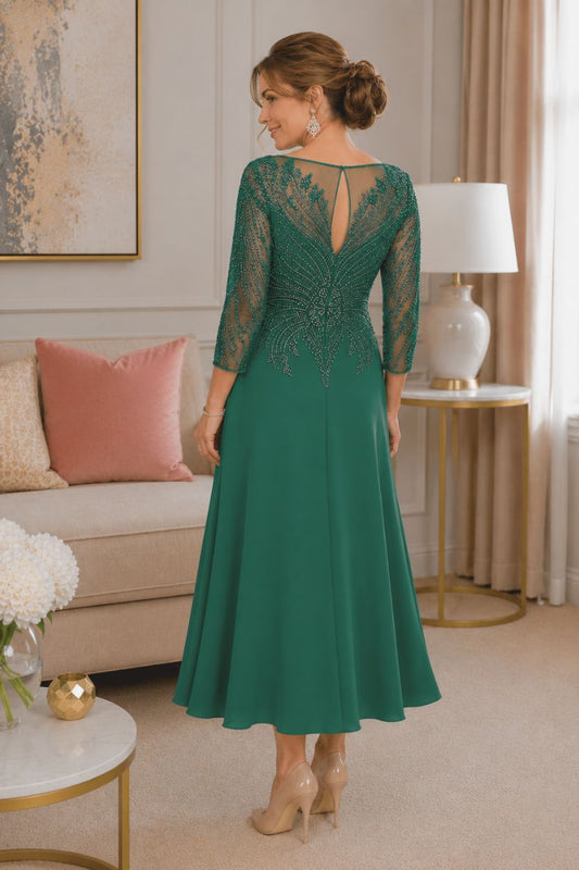 Back view of emerald green tea-length mother of the bride dress featuring a sheer illusion keyhole back, detailed beadwork, three-quarter sleeves, and a flowing crepe skirt on a mature brunette model in a luxury indoor boutique setting.