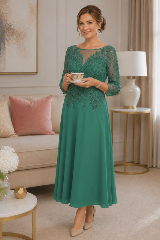 Emerald green tea-length mother of the bride dress with intricate beaded bodice, illusion neckline, three-quarter sleeves, and flowing crepe skirt on a mature brunette model in an elegant indoor boutique setting.