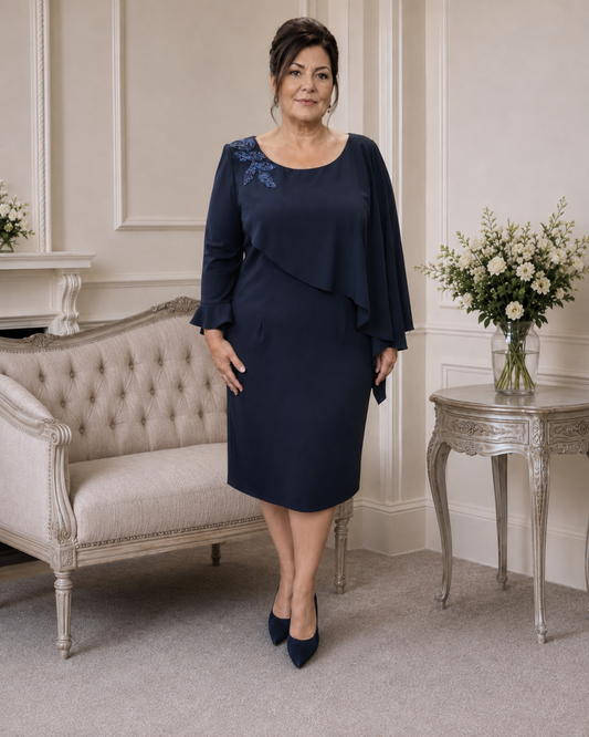 A woman stands in a formal indoor room wearing a navy knee-length mother of the bride dress with three-quarter sleeves, a draped asymmetrical overlay, and blue floral embroidery on the upper left shoulder. She is posed beside a tufted sofa and a decorative side table with flowers.