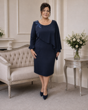A woman stands in a formal indoor room wearing a navy knee-length mother of the bride dress with three-quarter sleeves, a draped asymmetrical overlay, and blue floral embroidery on the upper left shoulder. She is posed beside a tufted sofa and a decorative side table with flowers.